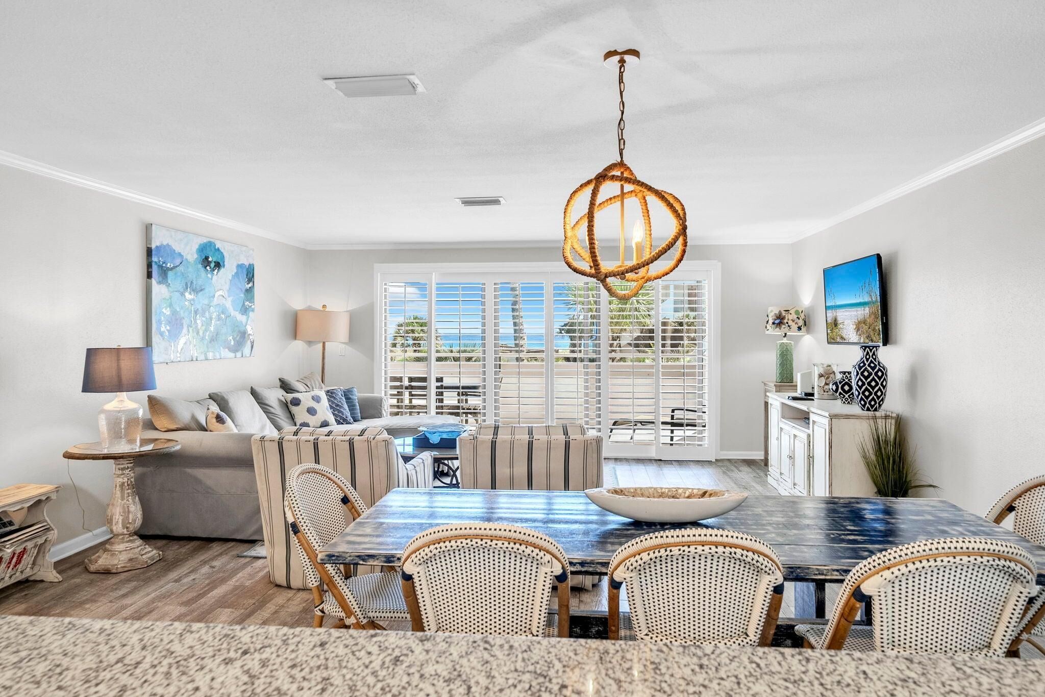 A dining area with a large table, wicker chairs, a hanging light fixture, and a view through large windows.