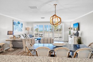 A dining area with a large table, wicker chairs, a hanging light fixture, and a view through large windows. - Aegean 210 (Destin)