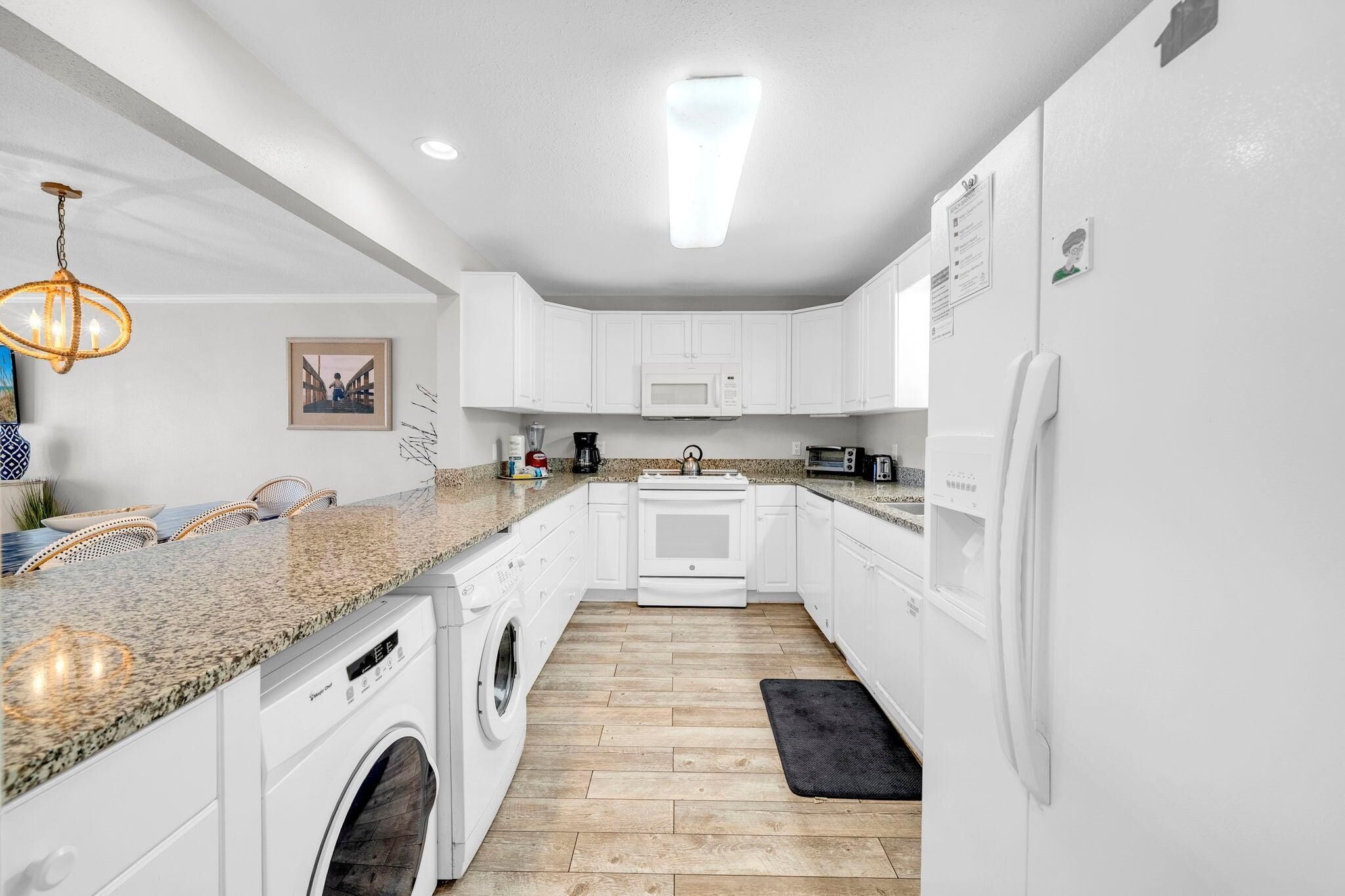 A modern kitchen with white cabinets, a granite countertop, a washing machine, a microwave, and a refrigerator.