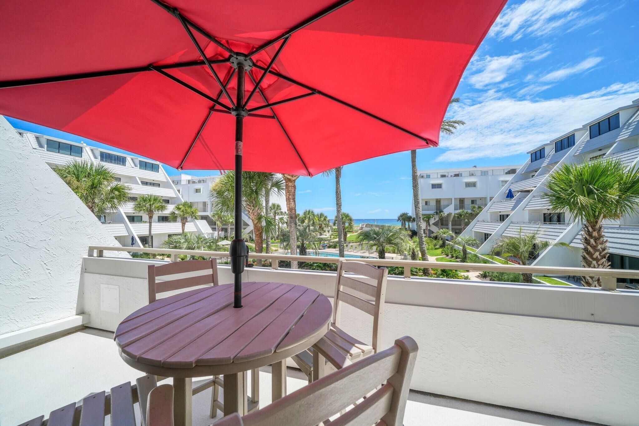 A balcony with a red umbrella, wooden table, and chairs, overlooking a pool and buildings.