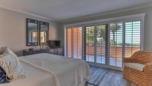 A bedroom with a bed, a TV, a dresser, a chair, and a balcony with a view of palm trees. - Aegean 210 (Destin)