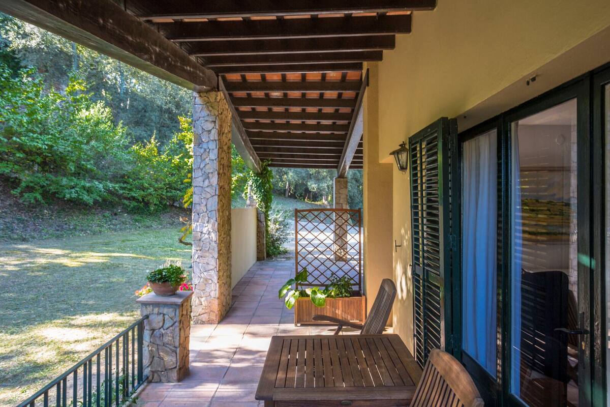 Un porche techado con una mesa y sillas de madera, pilares de piedra y vista a un jardín.