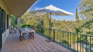 Un balcón con una mesa lista para comer, un paraguas grande y vistas a una zona boscosa.