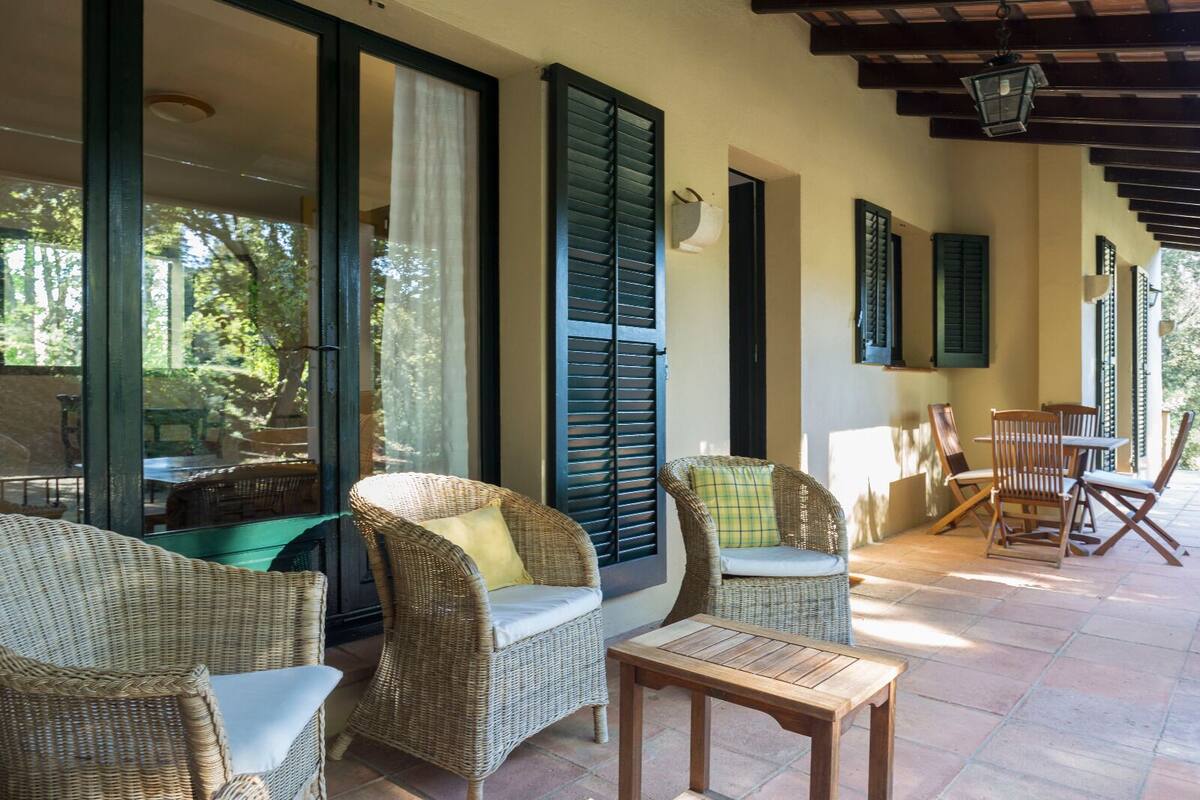 Un espacio de estar al aire libre cubierto con mobiliario de mimbre y una mesa de madera, con amplias ventanas y contraventanas.