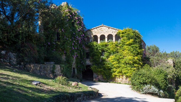 Un edificio histórico de piedra con arcos, rodeado de abundante vegetación y un sendero de grava que conduce hacia él.