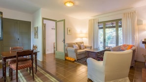 Una sala de estar con una mesa de comedor, dos sofás y una mesa de centro. Hay una puerta que conduce a otra habitación y una ventana con cortinas.