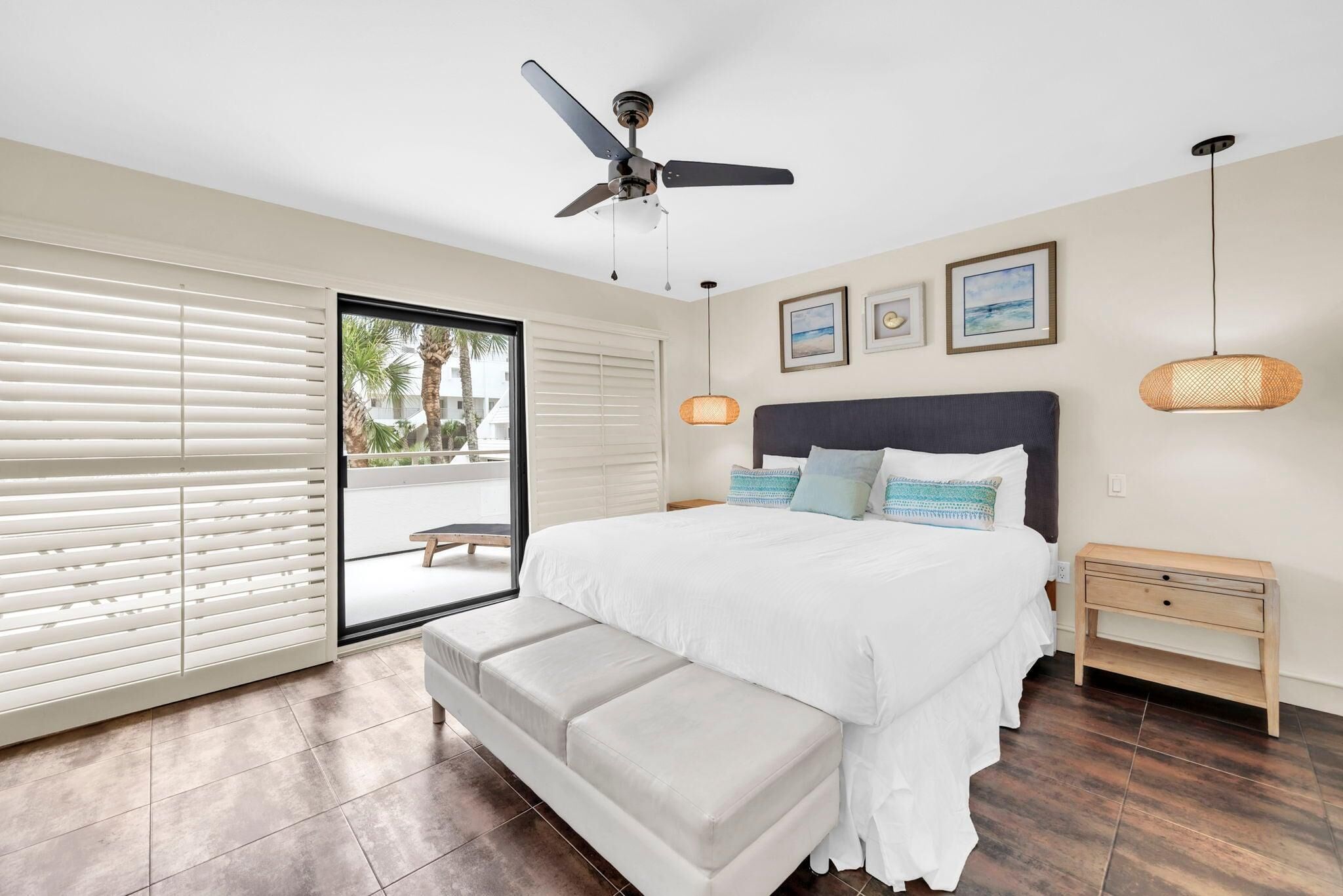 A bedroom with a bed, a ceiling fan, a bench, a nightstand, and a view through a glass door.
