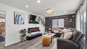A modern living room with a grey sofa, a flat-screen TV, and a chandelier. - Super Spacious Family Vacation Home! (San Diego)