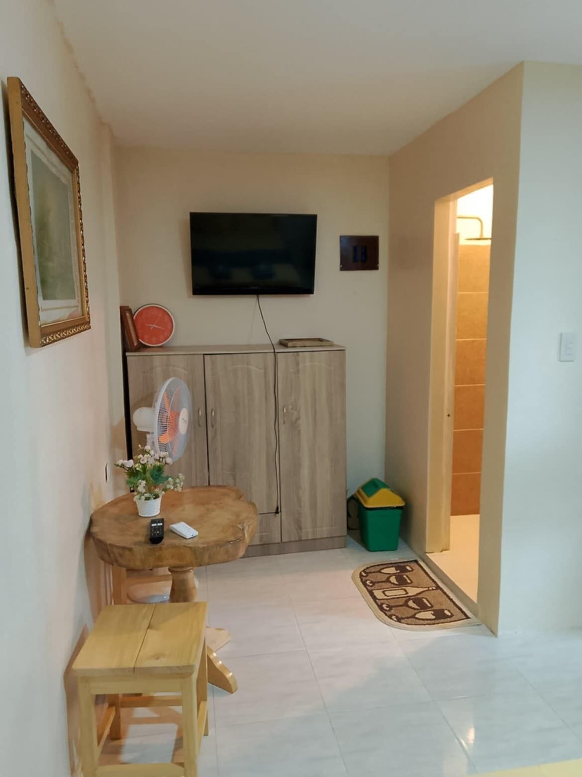 Una sala de estar con un televisor montado en la pared, un mueble de madera, una mesita con un jarrón de flores y un cubo de basura verde.