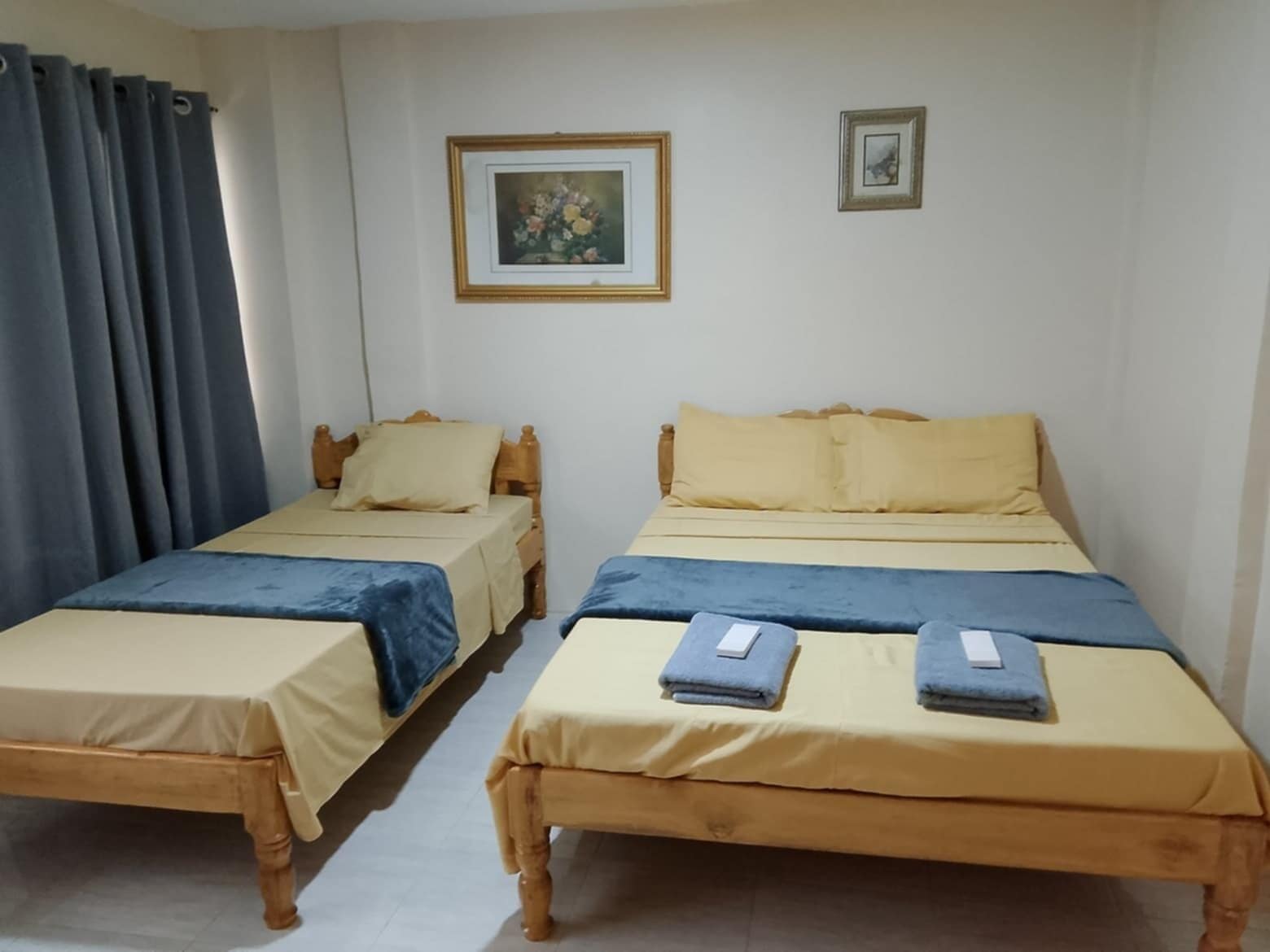 A room with two single beds, each with a wooden frame and a blue bedspread. There are two pillows on each bed, one beige and one yellow. A framed picture hangs on the wall above the beds.