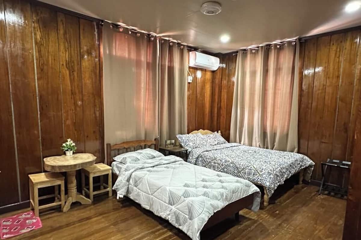 A room with two beds, a small round table with a chair, a wooden floor, and an air conditioning unit on the wall.