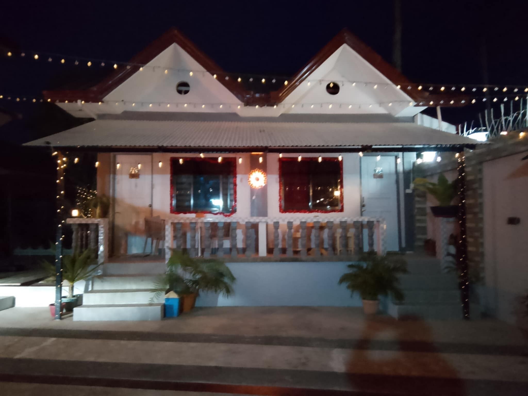 A two-story building with a white facade and a red roof. There are two visible windows on the front, one with a red frame and one with a white frame. The building is decorated with string lights.