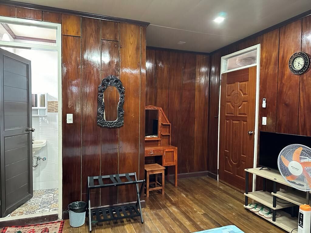 Une pièce aux murs en bois, avec une porte en bois, un miroir, une petite table en bois, une chaise, un ventilateur et une horloge au mur.