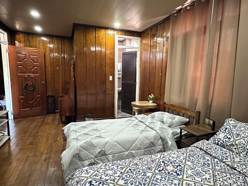 A room with wooden paneling, a bed with a patterned bedspread, a small table with a vase, and a door with a wreath.