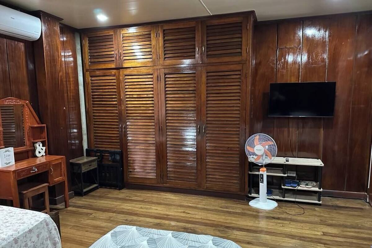 A room with a wooden wardrobe, a bed, a desk, and a TV mounted on the wall.