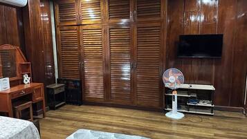 A room with a wooden wardrobe, a bed, a desk, and a TV mounted on the wall.