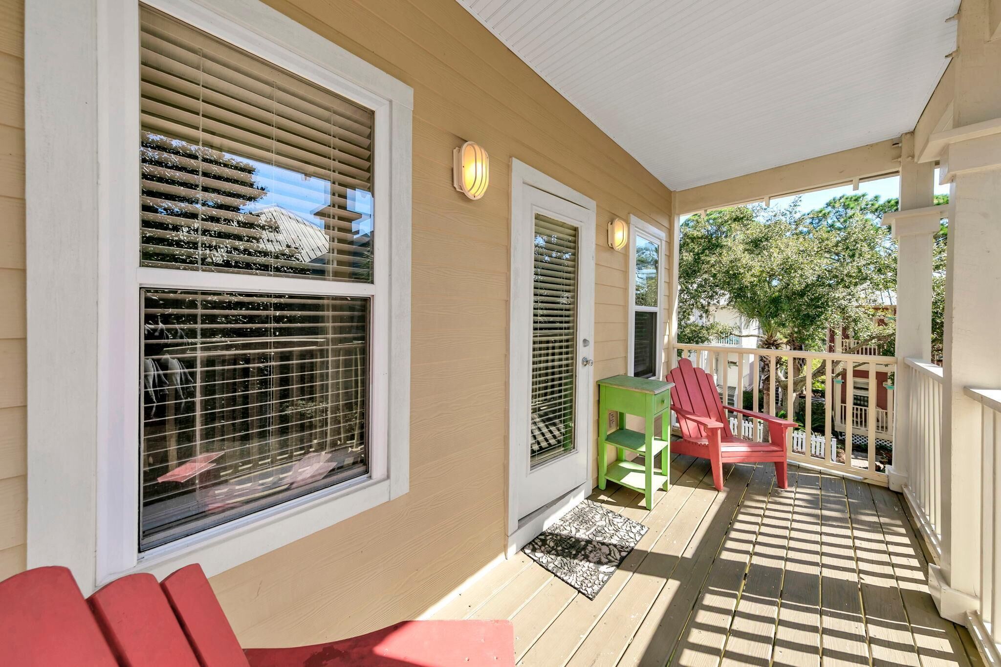 A covered porch with a wooden floor, a red chair, a green chair, and a white door.