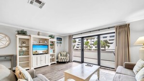 A living room with a TV, sofa, coffee table, and a view of a building through the window. - Aegean 213 (Destin)