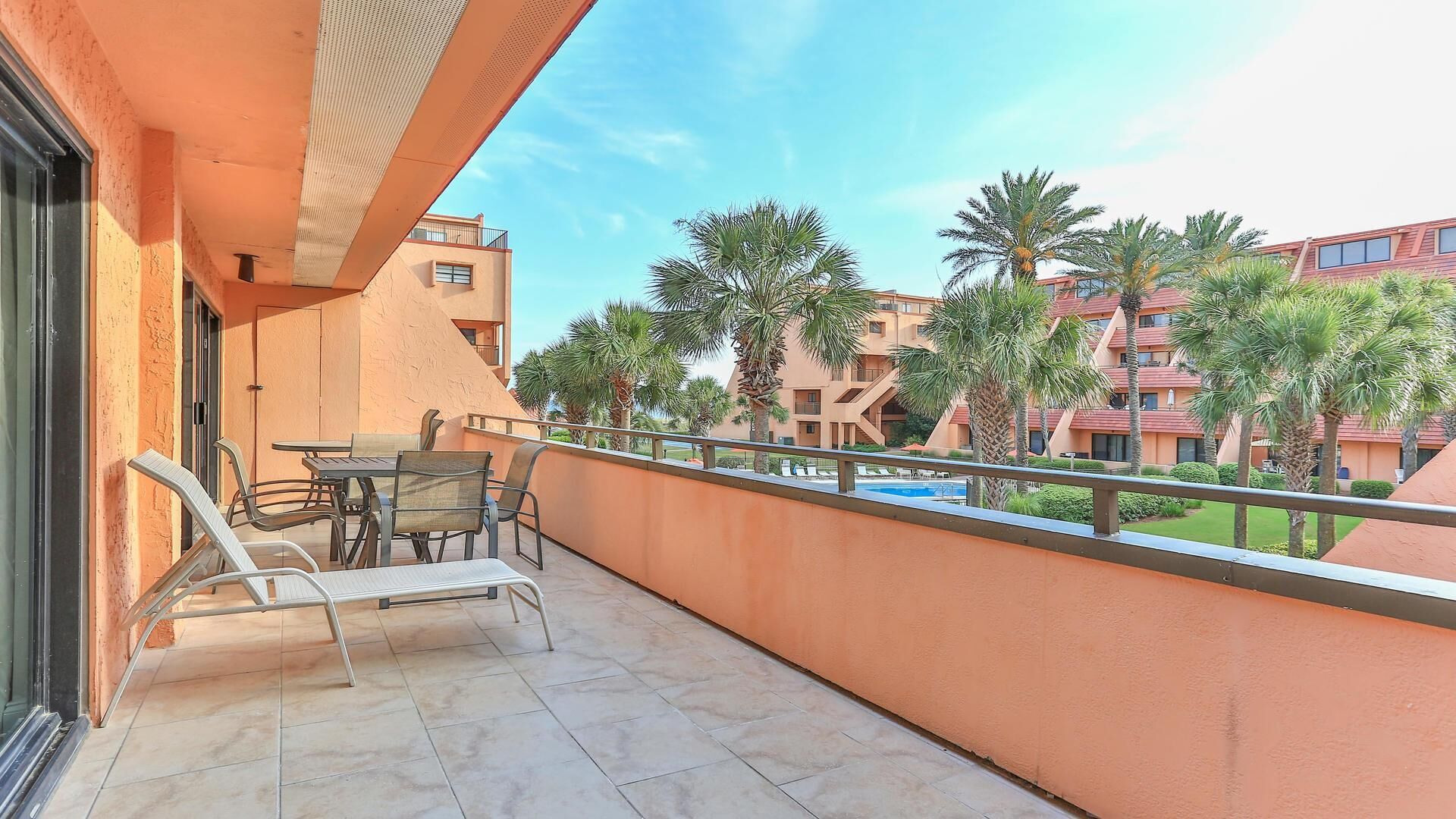 A balcony with a table and chairs, overlooking a pool and palm trees, with a building in the background.