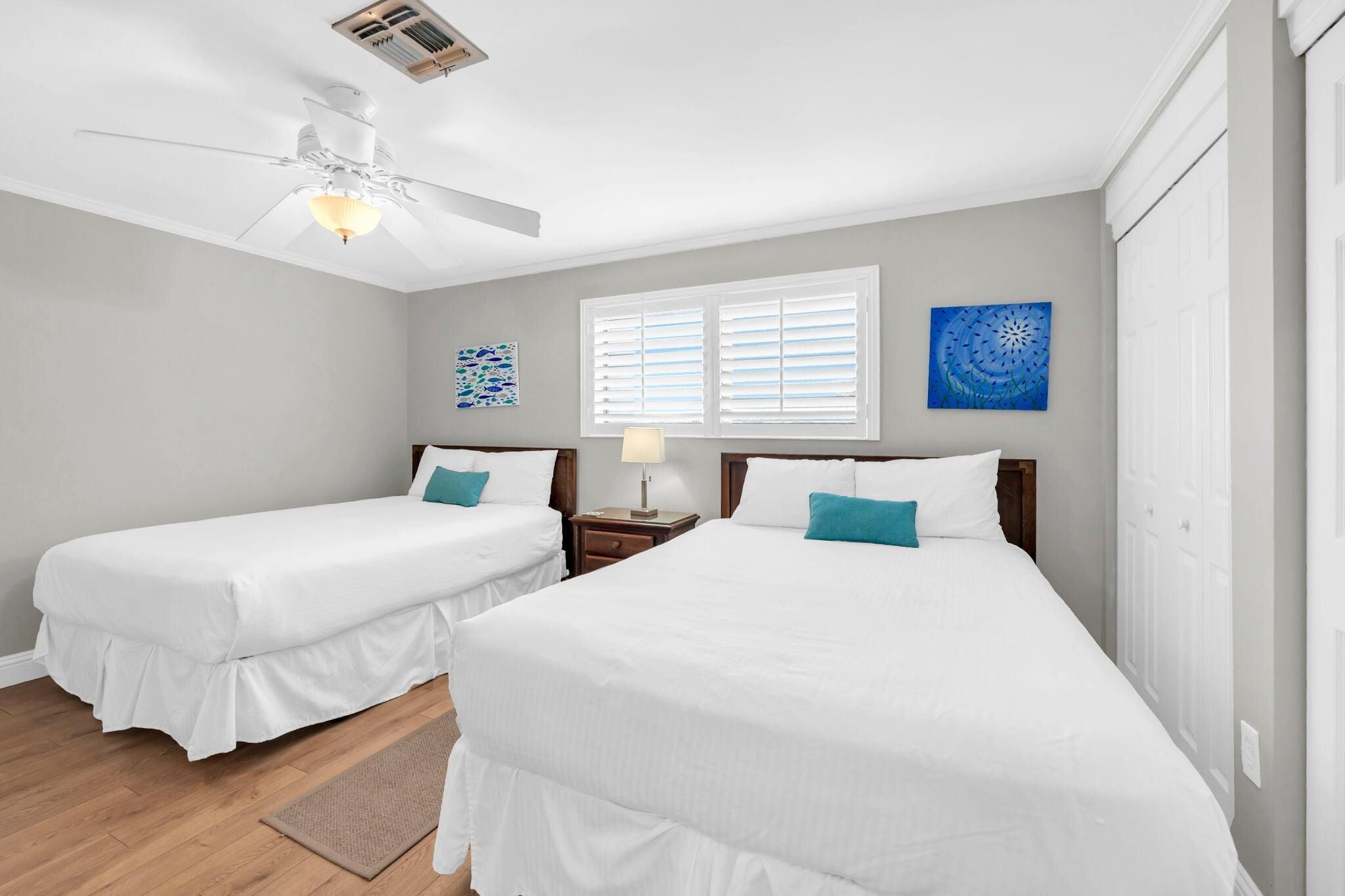 A hotel room with two beds, a ceiling fan, a window with blinds, and artwork on the walls.