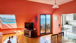 Sala de estar com televisão, mesa de centro de vidro e vista para o mar através de uma grande janela.