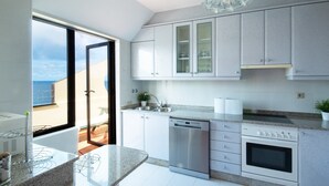 Cozinha moderna com máquina de lavar louça, forno e eletrodomésticos de aço inoxidável. Há uma janela grande com vista para o mar.