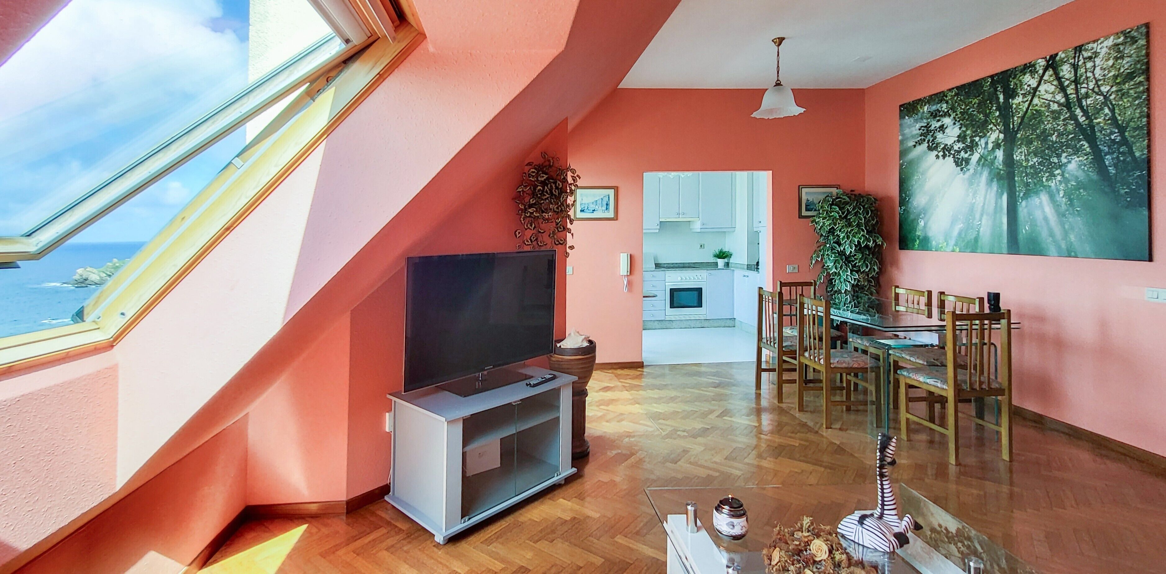 Een kamer met een groot raam, een flatscreen televisie, een eettafel met stoelen en een keukenruimte zichtbaar door een geopende deur.