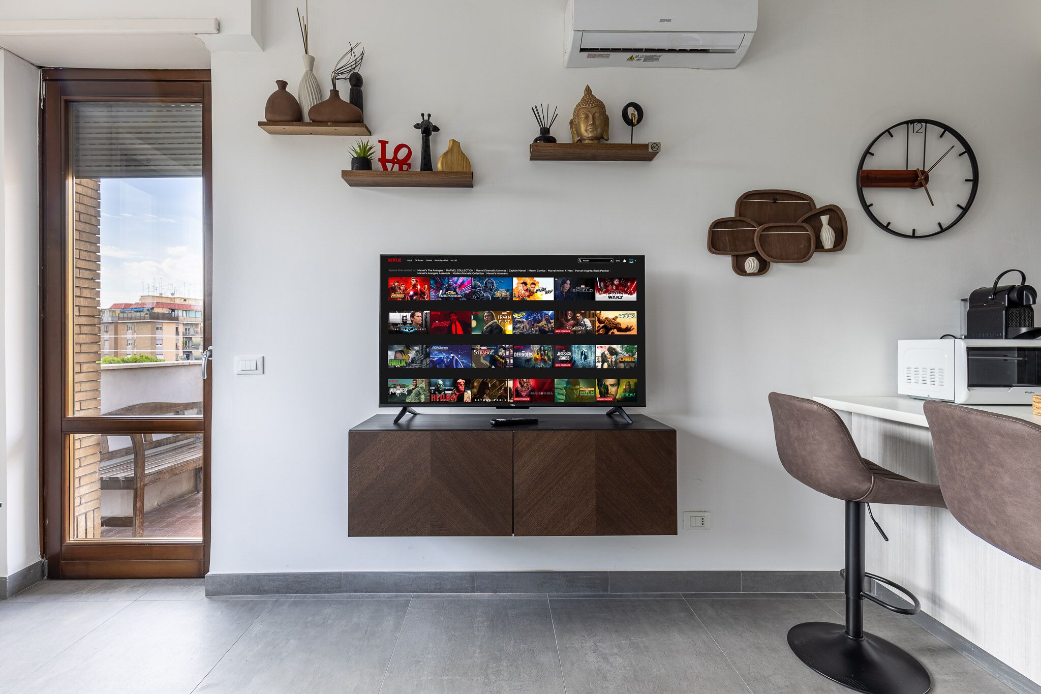 Een moderne woonkamer met een flatscreen televisie bevestigd aan een houten kast, een wandklok en een keukenhoek met een magnetron.