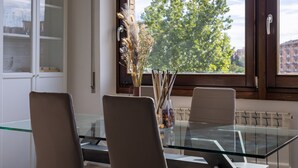 Un comedor con una mesa de vidrio y sillas, un jarrón con plantas secas y una ventana con vistas a árboles y edificios.