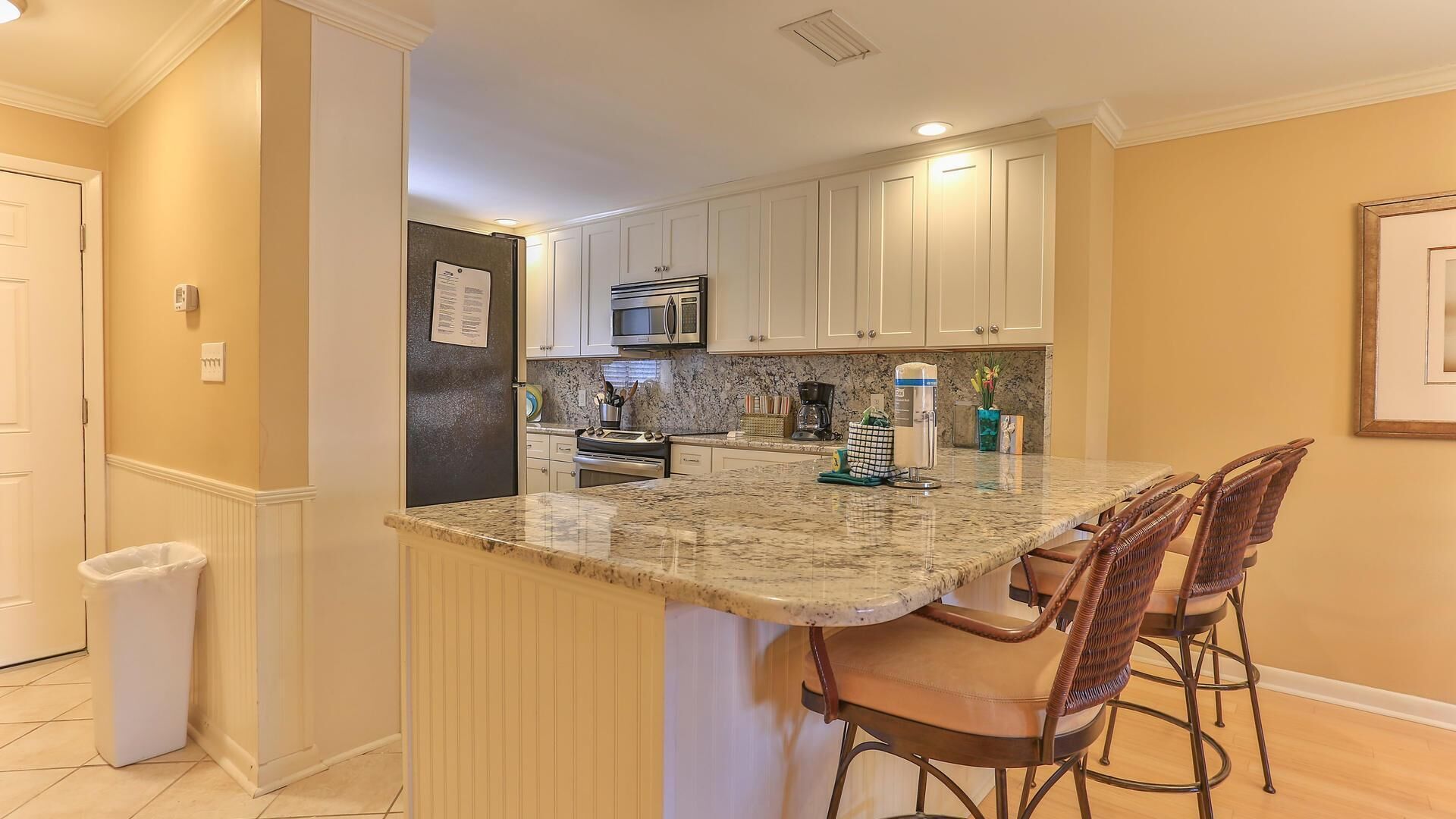 A kitchen with a granite countertop, white cabinets, a stainless steel refrigerator, and a microwave. There are two bar stools and a trash can.