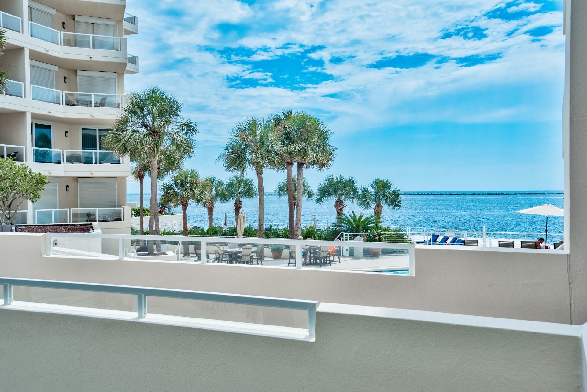 A balcony with a view of the sea, palm trees, and outdoor seating.