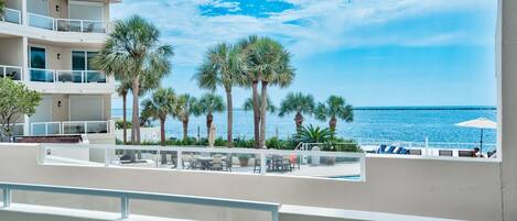 A balcony with a view of the sea, palm trees, and outdoor seating.