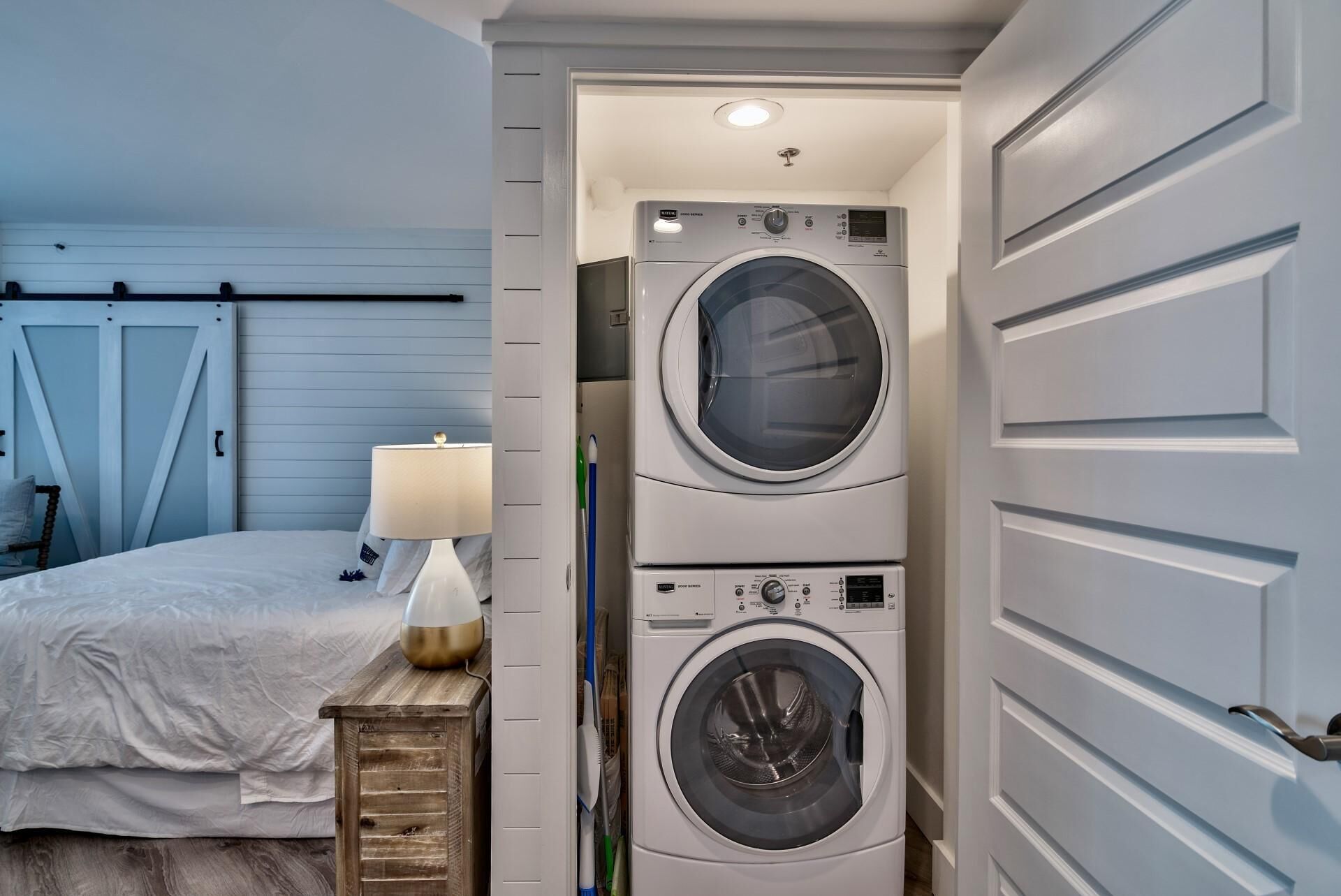 A laundry room with two stacked front-loading washing machines, a bed with white bedding, a wooden nightstand with a lamp, and a door with a handle.