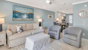 A living room with a sofa, two armchairs, a coffee table, a painting, and a dining area with a table and chairs. - Poolside Villas 106 (Destin)