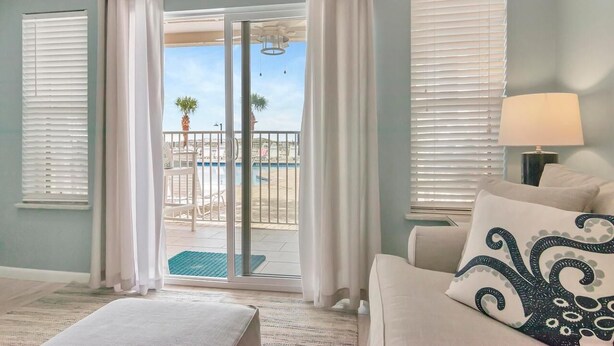 A room with a sliding glass door leading to a balcony with a pool, white curtains, a beige sofa with a decorative pillow, and a lamp on a side table. - Poolside Villas 106 (Destin)