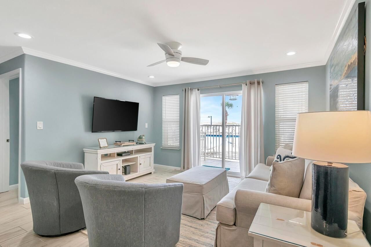 A living room with a flat-screen TV, grey sofas, a coffee table, a ceiling fan, and a balcony with a view.