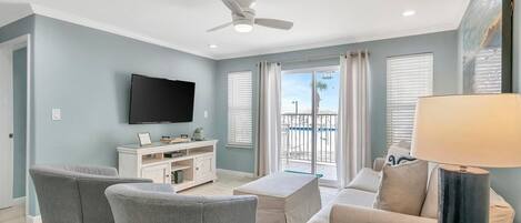 A living room with a flat-screen TV, grey sofas, a coffee table, a ceiling fan, and a balcony with a view.