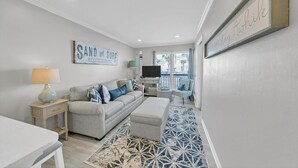Ett modernt vardagsrum med en grå soffa, en tv och en skylt med texten ”SAND and SURF DESTIN, FLORIDA”.