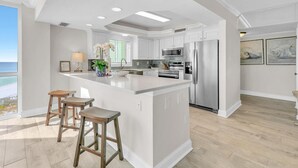 A modern kitchen with a central island, white cabinetry, stainless steel appliances, and a view of the ocean. - Shoreline Towers 1061 (Destin)