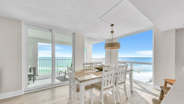 A dining area with a view of the ocean, white chairs, a wooden table, and a hanging light fixture. - Shoreline Towers 1061 (Destin)