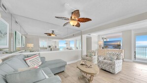 A spacious living room with a grey sofa, patterned armchair, and a wooden coffee table. A ceiling fan with leaf designs is visible. - Shoreline Towers 1061 (Destin)