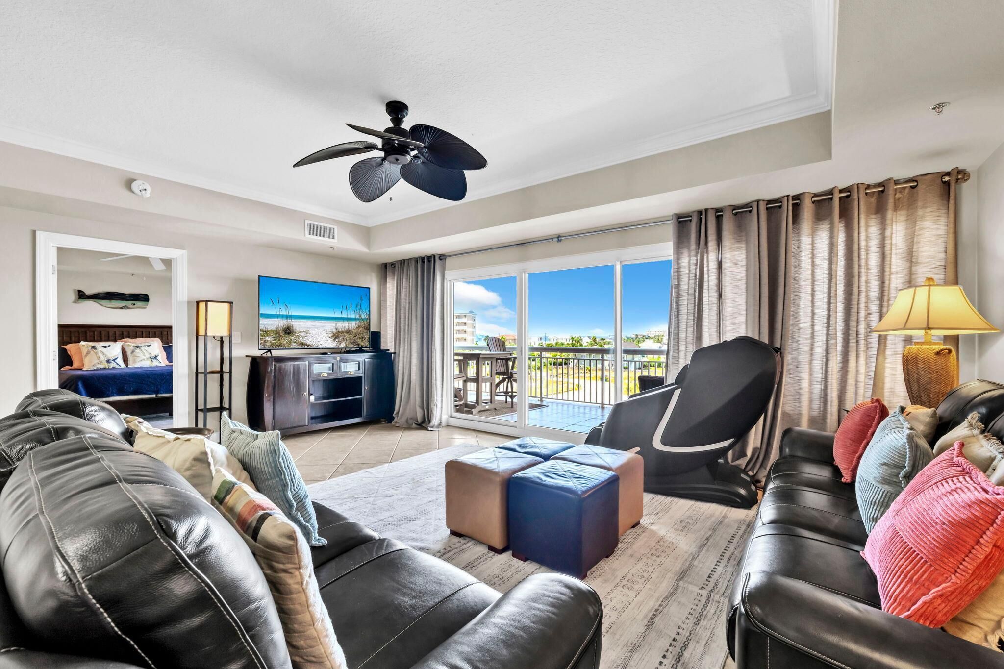 A spacious living room with a large window, a ceiling fan, a flat-screen TV, and a balcony with outdoor furniture.