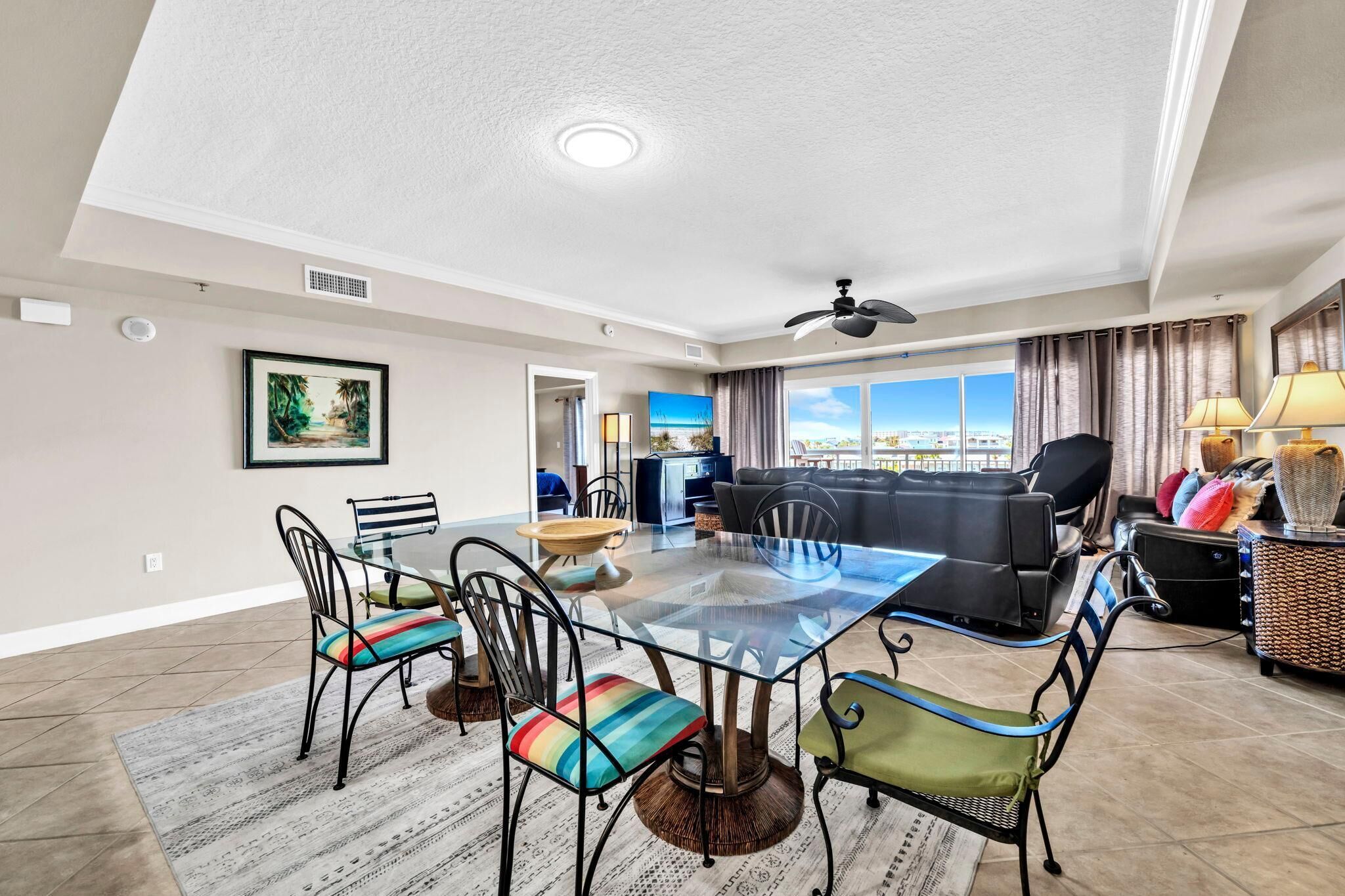 A spacious living room with a glass-top dining table, comfortable seating, a ceiling fan, and a large window offering a view.