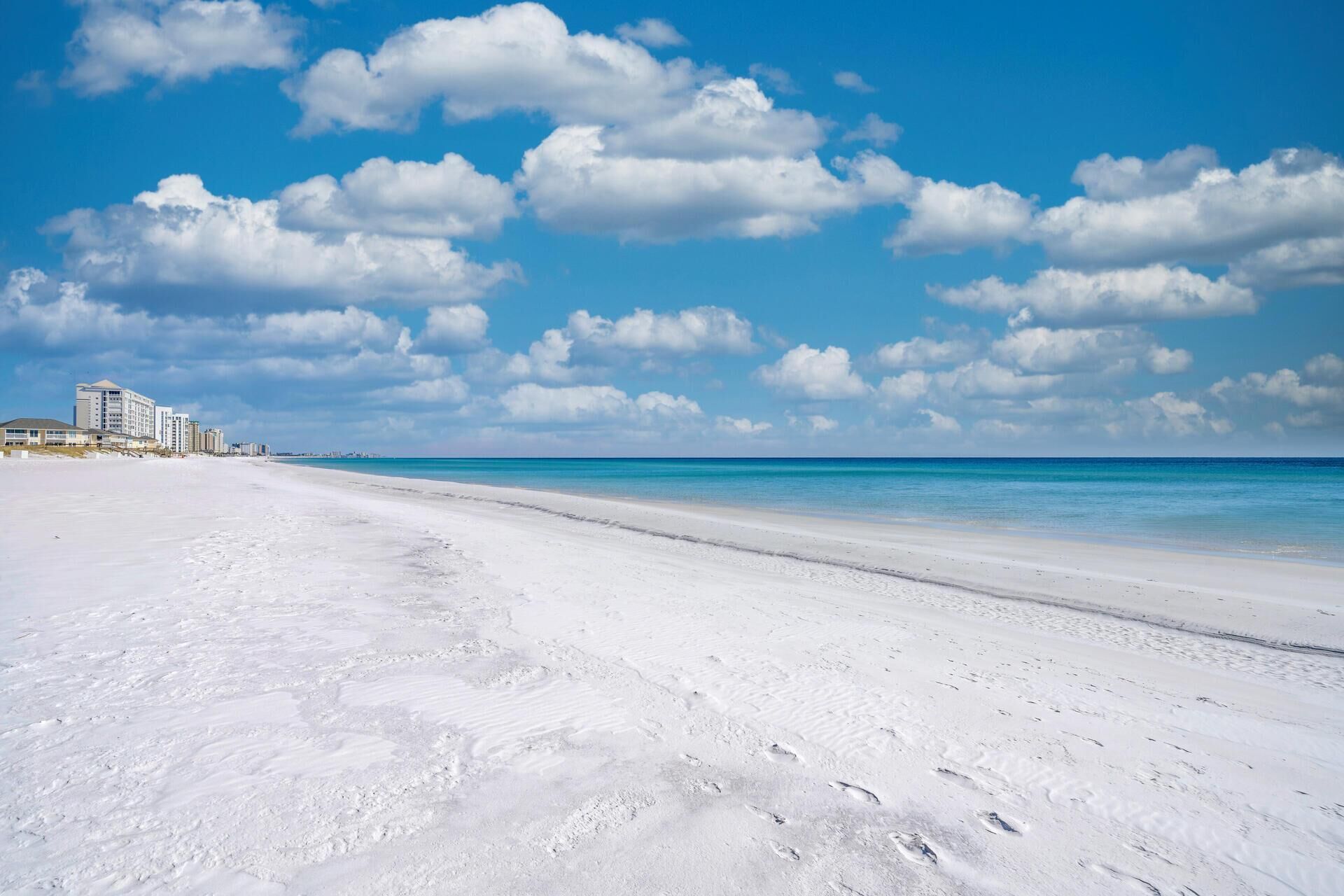 A beach with white sand and a clear blue sky. There are buildings in the background.