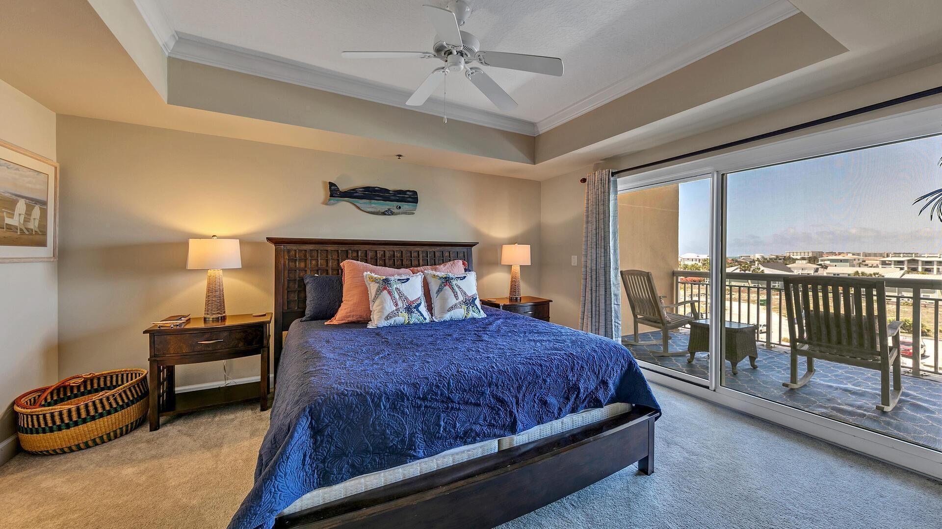 A bedroom with a large bed, a ceiling fan, a nightstand, a lamp, a wicker basket, and a balcony with a view.