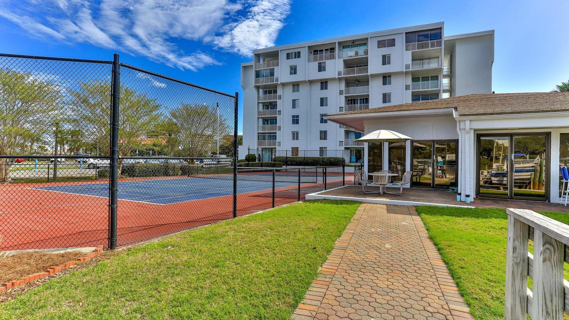 Una cancha de tenis rodeada por una cerca de malla metálica, un área de descanso cubierta con mesas y un edificio de varios pisos al fondo.