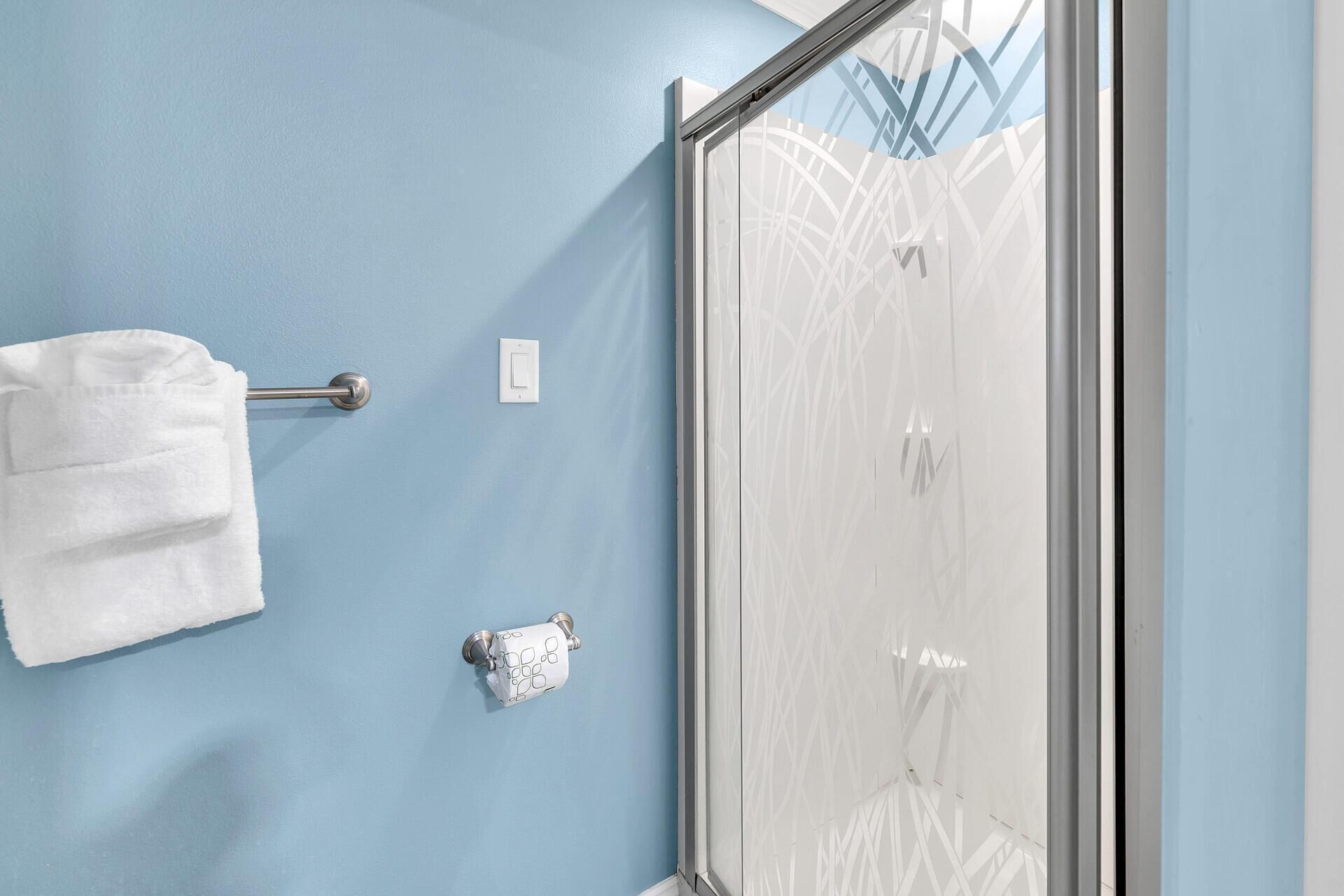 Un baño con pared azul, una toalla blanca en un perchero, un dispensador de papel higiénico y una puerta de ducha de cristal.