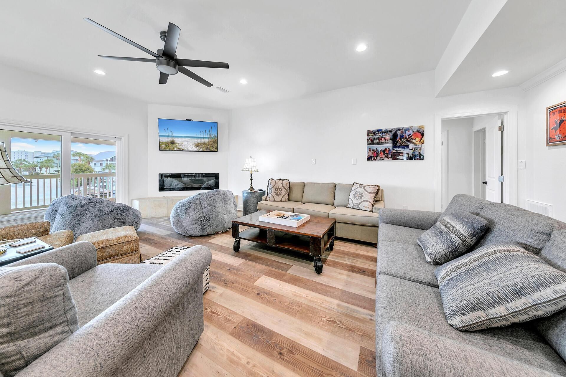 Un salon spacieux avec un grand canapé, une table basse et un ventilateur de plafond. Un téléviseur est fixé au mur et une fenêtre offre une vue sur la plage.