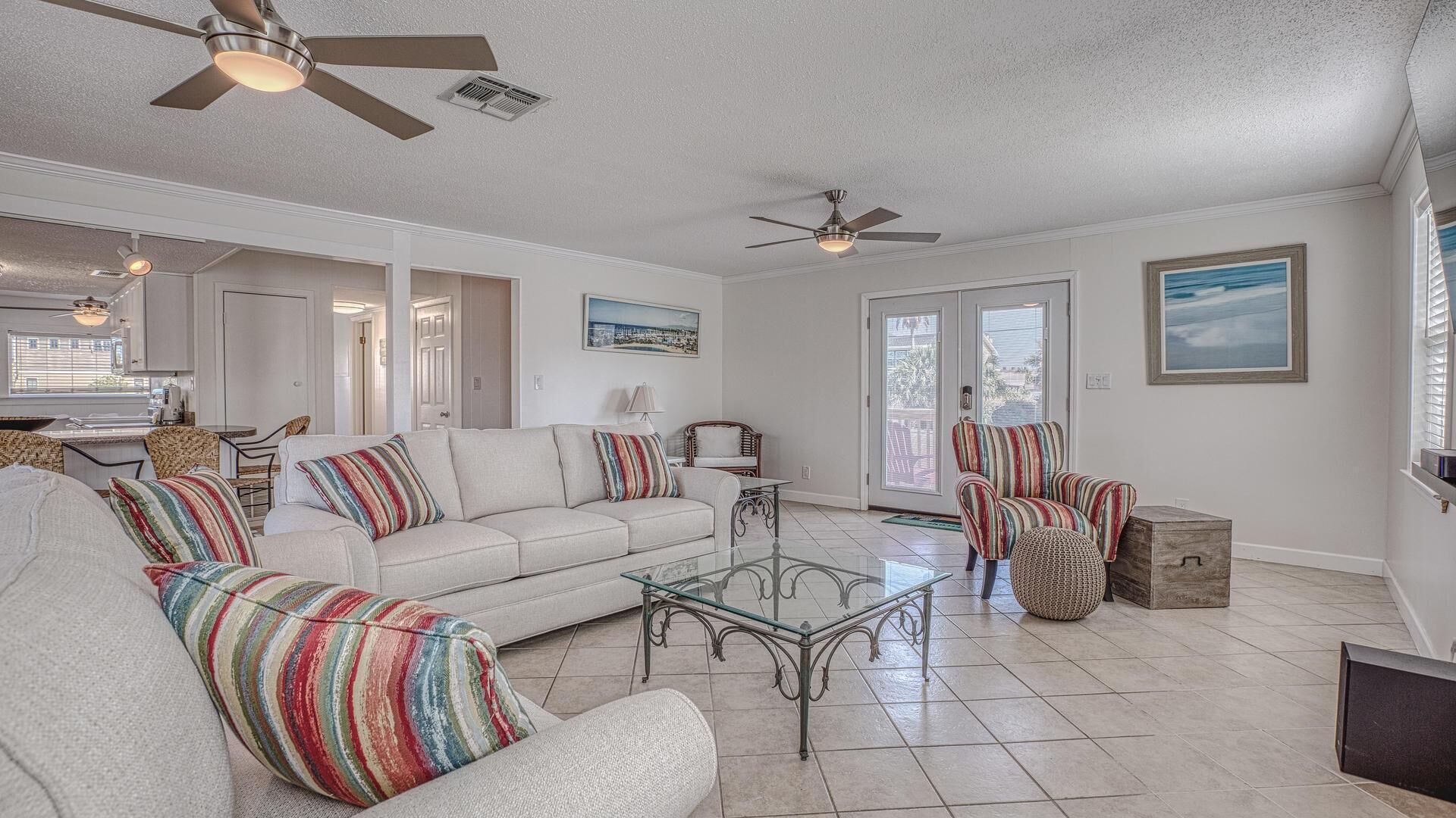 A spacious living room with a sofa, armchairs, a coffee table, and a dining area with a table and chairs. There are ceiling fans and a large window with blinds.