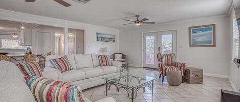 A spacious living room with a sofa, armchairs, a coffee table, and a dining area with a table and chairs. There are ceiling fans and a large window with blinds.